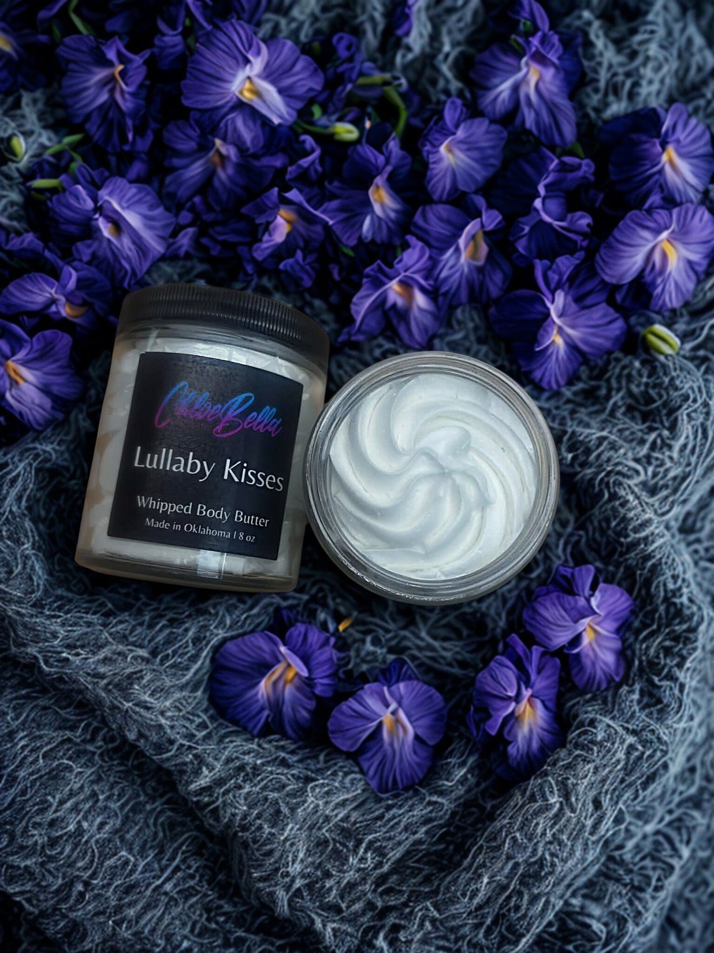 Whipped body butter jar labeled 'Lullaby Kisses' surrounded by purple flowers on a textured gray fabric background.